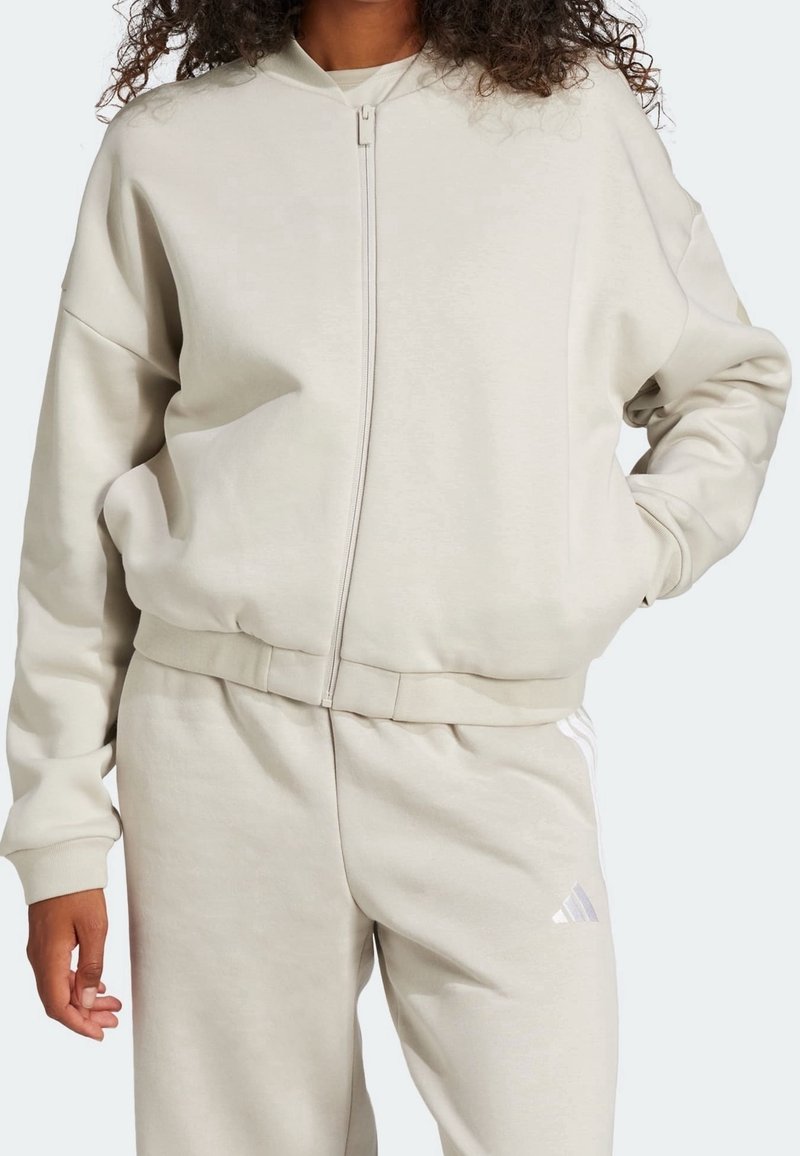 Person som bär en ljusbeige zip-up hoodie och matchande sweatpants med en liten Adidas-logga på låret, med en hand i fickan.