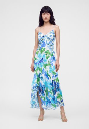 Vrouw met lang zwart haar die een witte mouwloze jurk met blauw-groene bloemenprint draagt en gouden open teen hakken, staand op een effen achtergrond.