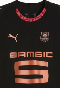 Juoda sportinė marškinė su apvaliu kaklu, su raudonu raštuotu kaklu, vario spalvos logotipais ir dideliu užrašu "SAMSIC" priekyje.