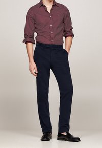 Marineblauwe chinos gecombineerd met een bordeauxrood, gestreept overhemd met knopen. Het overhemd heeft kleine witte stippen en opgerolde mouwen. Zwarte schoenen maken de outfit compleet.