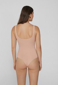 Žena stojící zády k fotoaparátu, oblečená v světle růžovém bezrúškovém body, na jednobarevném šedém pozadí.