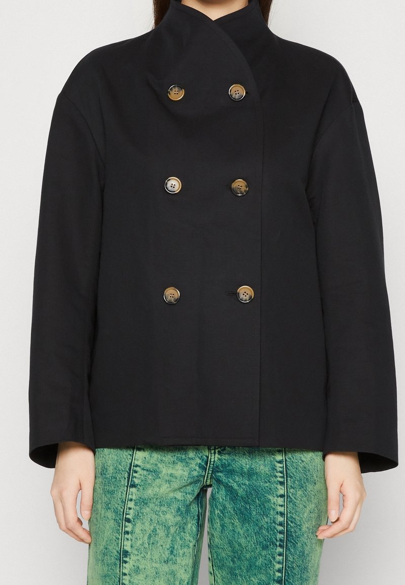 Zwarte double-breasted jas met een hoge kraag, gemaakt van een gladde stof. Voorzien van gouden knopen en brede mouwen, gecombineerd met groene denim.