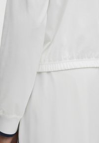 Valge jakk pehme tekstuuriga, mis on varustatud volditud hemiga ja tumesiniste aktsentidega mansettides. Puhas, minimaalne disain.