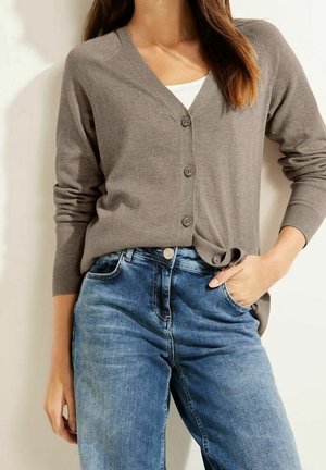 Kobieta w brązowym kardiganie zapinanym na guziki, nosząca biały top i niebieskie dżinsy, stojąca z jedną ręką w kieszeni na jednolitym tle.