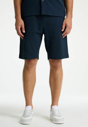 Man met donkere textuurschakel shorts, witte sokken en witte sneakers, staand tegen een effen lichte achtergrond.