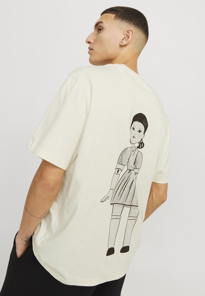 Camiseta de algodón color crema con mangas cortas que presenta una ilustración negra de una muñeca en la parte de atrás. Corte relajado y textura suave.