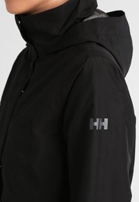 Black waterproof jacket with a high neck and hood. Features a silver logo on the left sleeve and a textured inner lining.