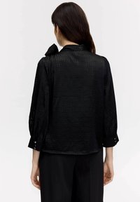 Blusa nera con un motivo testurizzato, colletto pieghettato e polsini con bottoni. Maniche lunghe voluminose, tessuto leggero e vestibilità comoda.
