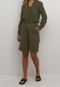 Femme portant un blouse à manches longues vert olive et un short cargo assorti jusqu'aux genoux avec des poches, associé à des sandales à glissière noires.