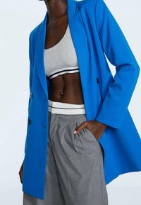 Blazer bleu croisé à double boutonnage avec une texture lisse, porté sur un haut court gris à rayures noires et blanches, assorti à un pantalon plissé gris.