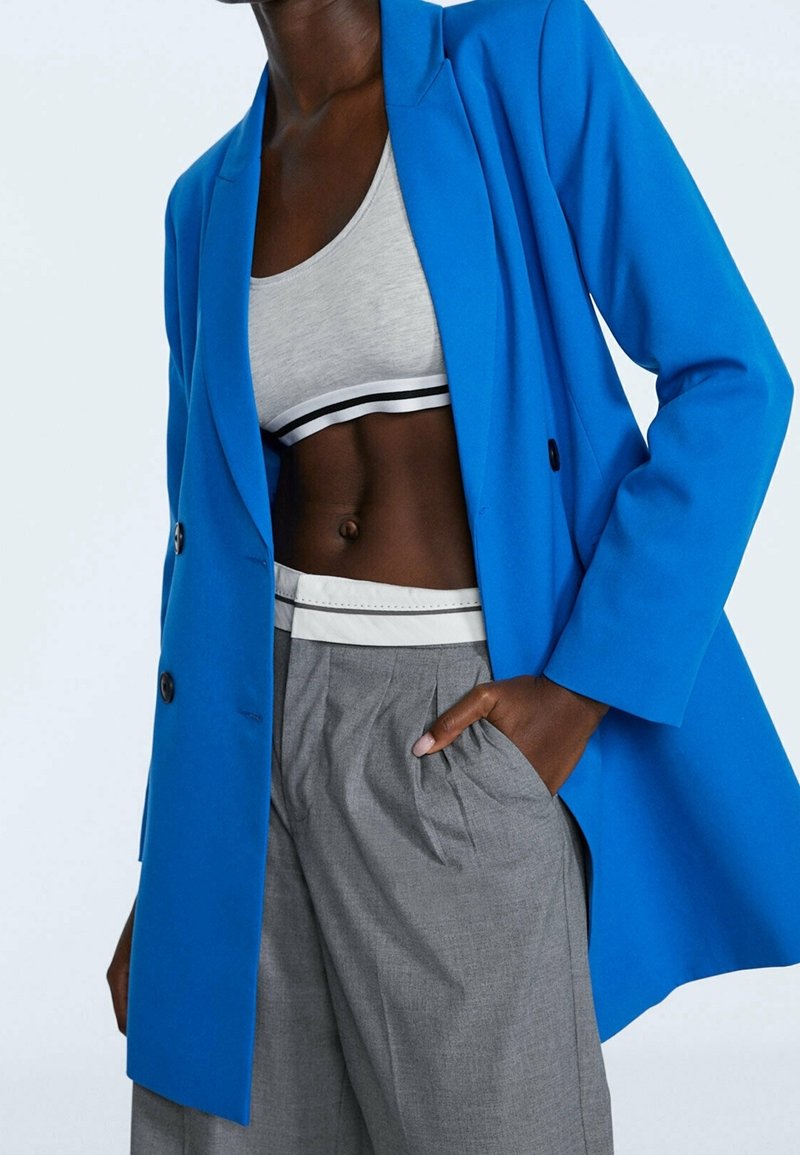 Blazer bleu croisé à double boutonnage avec une texture lisse, porté sur un haut court gris à rayures noires et blanches, assorti à un pantalon plissé gris.