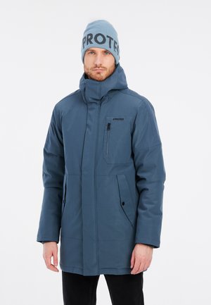 Blauwe geïsoleerde jas met hoge kraag, voorzakken en een ritszak op de borst. G gedragen met een lichtblauwe beanie met donkere tekst.