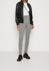 Svart faux läder bomberjacka, vit t-shirt med rund hals, byxor i svart och vitt hundtandmönster, svarta sneakers, stående mot en vit bakgrund.