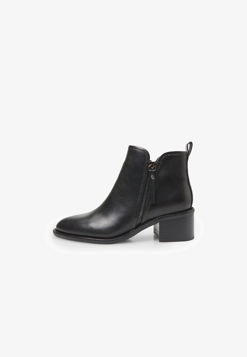Bota de tobillo de cuero negro con cierre de cremallera lateral, punta redondeada y tacón grueso. Presenta un tirador en forma de lazo en la parte trasera.
