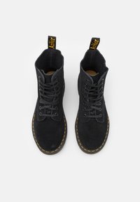Botas de tobillo de ante negro con costuras amarillas, de punta redonda y cordones de seis ojales. Presentan un tirador de tela en la parte trasera. Superficie texturizada.