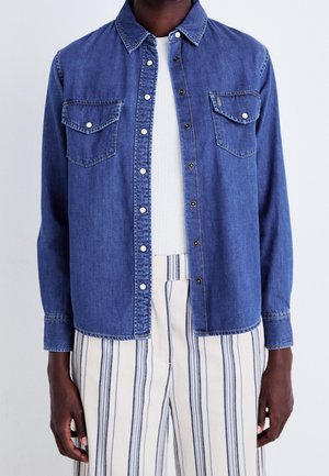Person wearing an unbuttoned blue denim shirt over a white top with cream pants featuring vertical blue stripes.