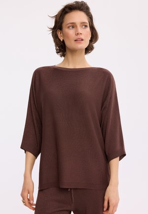 Kobieta ubrana w luźny, brązowy sweter z rękawami do łokci i pasujące spodnie, lekko patrząca na bok na jednolitym tle.