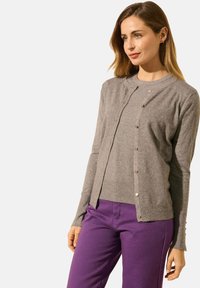 Cardigan gris clair à manches longues, avec un col rond, une fermeture à boutons et une texture douce. Associé à un pantalon violet.