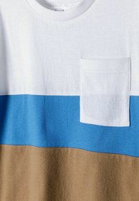 Medvilninis marškinėliai su horizontaliomis baltos, mėlynos ir rudos spalvos juostelėmis. Įtraukta maža balta krūtinės kišenė ir apvalus kaklas.