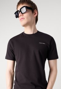 Fekete pamut póló rövid ujjakkal, az elülső részen egy kis fehér "Calvin Klein" logóval. A modellen túlnyúló fekete napszemüveg van.