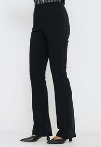 Donna che indossa pantaloni neri a vita alta aderenti e tacchi a punta neri con tacchi a forma di palla, in piedi su uno sfondo bianco semplice.