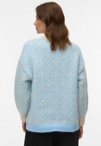 Femme aux cheveux bruns mi-longs portant un pull duveteux à motifs bleu clair et crème, vue de dos sur un fond blanc.