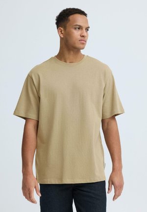 Jonge man die een effen beige oversized T-shirt en donkere broek draagt, staand en zijwaarts kijkend tegen een lichte achtergrond.