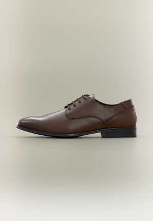 Chaussure habillée pour homme en cuir marron avec lacets et talon bas empilé sur fond beige uni.
