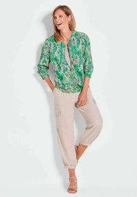 Groene bomberjack met bloemenpatroon en ritssluiting, gecombineerd met beige cargobroek met zakken en elastische manchetten. Schoeisel: tan sandalen met bandjes.