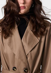 Beige dobbelspent trenchcoat med store svarte knapper, skarpe krager og glatt stofftekstur, båret over en svart ribbestrikket turtleneck.