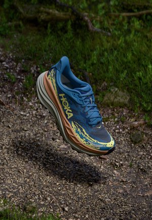 Zapatilla de running Hoka azul y amarilla con suela gruesa flotando sobre un fondo de sendero de grava y hierba.