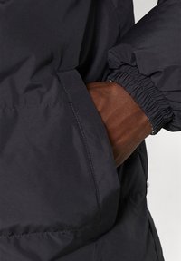 Zwarte pufferjack heeft een zijzak met elastische manchetten, en toont een gladde, matte textuur en subtiele stikseldetails.