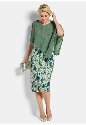 Vrouw in knie-lange bloemenjurk met groene doorschijnende cape, houdt een zilveren clutch vast en draagt zilveren hakken, staand tegen een witte achtergrond.