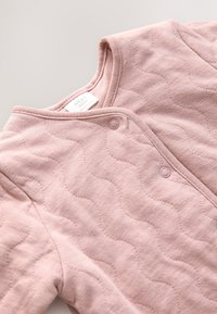 Vatteret babybeklædning i blødt pink stof. Har rundet halsudskæring og trykknapper. Tekstureret mønster med diskrete bølger på overfladen.