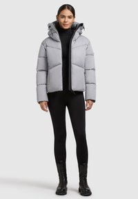 Giacca puffa grigia chiara con cappuccio, caratterizzata da sezioni trapuntate, chiusura a zip e una texture liscia. Indossata sopra leggings neri e stivali.