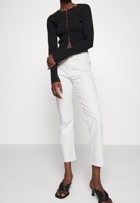 Femme portant un haut noir ajusté à manches longues avec des boutons, un jean blanc droit et des sandales noires à talons, debout devant un fond blanc.