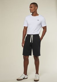 Weißes T-Shirt mit kurzen Ärmeln und kleinem dunklem Logo, kombiniert mit schwarzen Shorts mit Kordelzug. Getragen mit weißen und beigen Sneakers.