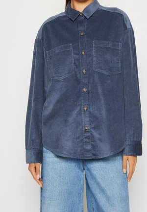 Persoon draagt een loszittend donkblauw corduroy overhemd met knopen en twee borstzakken, gecombineerd met lichtblauwe spijkerbroek.