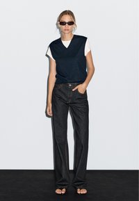 Gilet di maglia blu navy su una maglietta bianca, abbinato a jeans scuri a vita alta. Il modello indossa occhiali da sole neri e sandali minimalisti.