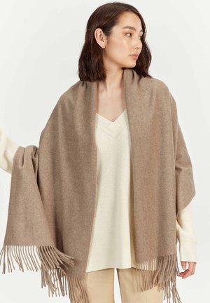 Woman wearing a beige shawl with fringe over a cream knit sweater and beige pants, looking to her right against a plain background.