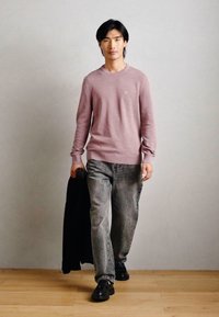 Lyserød tekstureret sweater, grå denimjeans, sorte sko og en sort jakke, med en glat lys baggrund og trægulv.