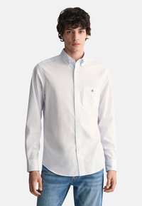 Chemise blanche boutonnée en tissu léger avec une poche poitrine, présentant une texture subtile et un emblème de marque sur la poche.