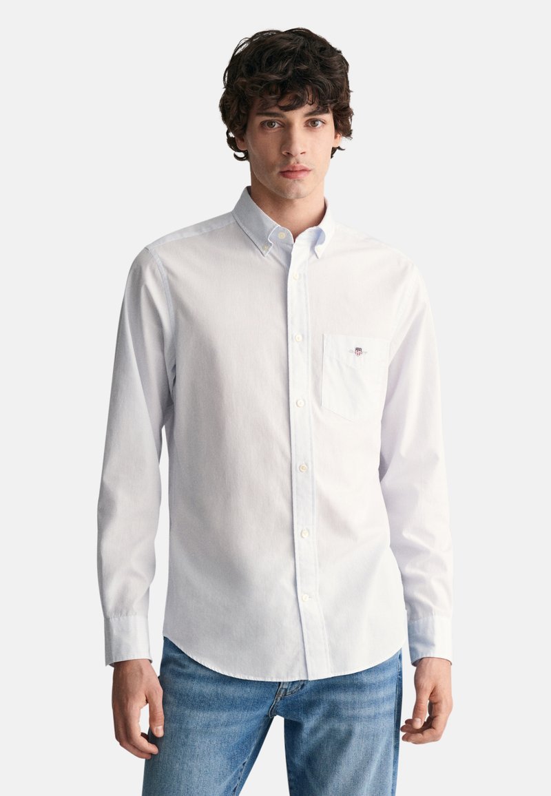 Chemise blanche boutonnée en tissu léger avec une poche poitrine, présentant une texture subtile et un emblème de marque sur la poche.