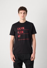 Czarny bawełniany T-shirt z wyraźnym czerwonym napisem: "CALVIN KLEIN NY" oraz "Calvin Klein Established 1968 NYC" w nowoczesnym designie.