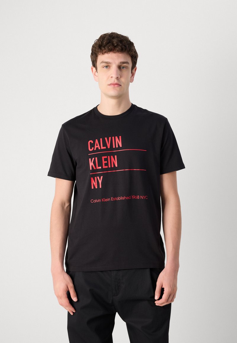 Camiseta de algodón negra con texto rojo en negrita: "CALVIN KLEIN NY" y "Calvin Klein Establecido 1968 NYC" en un diseño moderno.