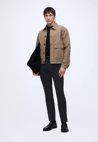 Uomo con giacca beige con bottoni, camicia nera, pantaloni neri e scarpe nere, che tiene una borsa nera su uno sfondo chiaro e uniforme.