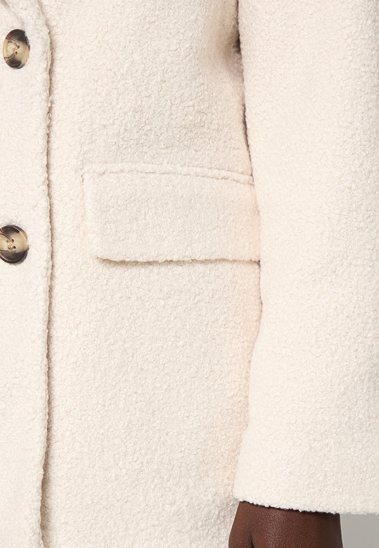 Kremowy, teksturowany płaszcz z zapięciem na guziki, kieszenią zapinaną na klapę oraz długim rękawem, z widoczną na mankiecie ciemną skórą.