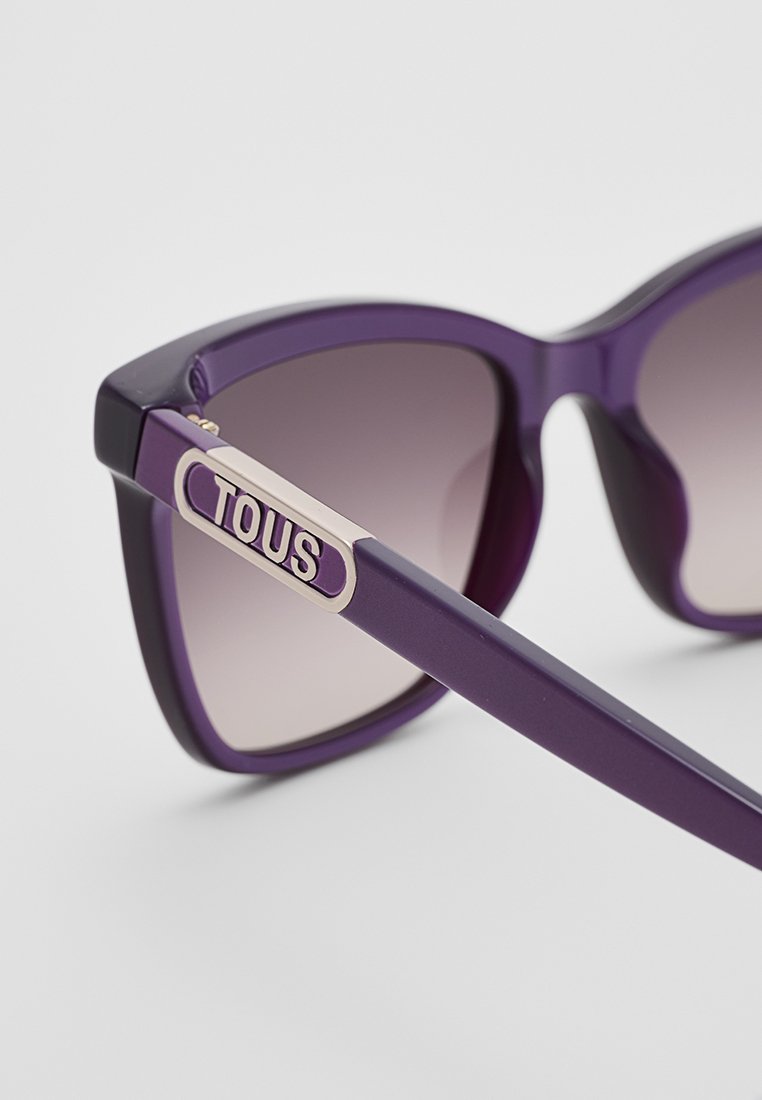 Fioletowe oversize okulary przeciwsłoneczne z gradientowymi szkłami. Charakteryzują się odważnym kocim kształtem i złotym akcentem z logo "TOUS" na ramieniu.