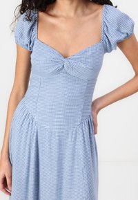 Blå och vit rutigt klänning med en åtsittande bodice, fyrkantig halsringning, puffärmar och en utsvängd kjol. Tillverkad av lätt tyg.