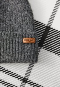 Gorro de malha cinza com textura canelada e um rótulo de couro castanho com "Barbour ENGLAND." O fundo apresenta um padrão xadrez em preto e branco.